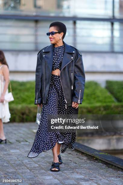 Tamu McPherson wears sunglasses, earrings, a black asymmetric flowing dress with white polka dots and a draped crop top, a black leather biker...