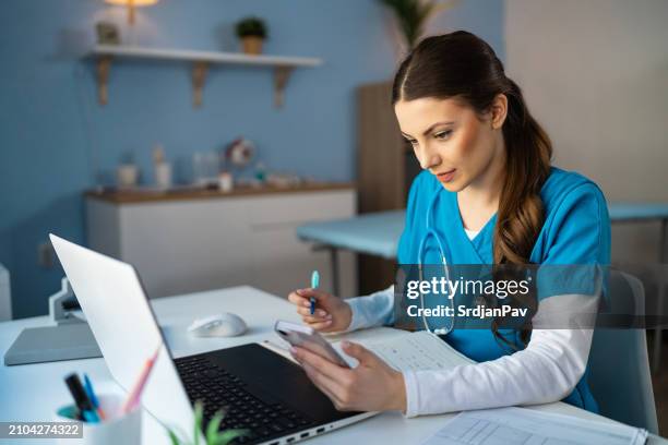 female doctor in blue medical scrubs, using mobile phone while working at the ordination - female doctor on computer stock pictures, royalty-free photos & images