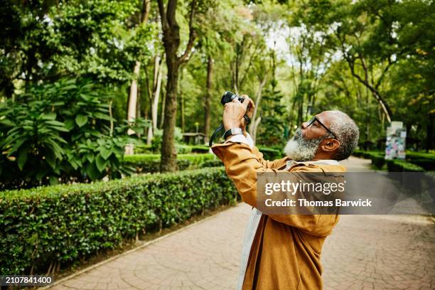 medium shot senior man taking photograph in park during city vacation - man taking photos stock pictures, royalty-free photos & images