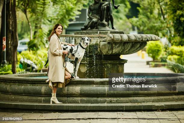 wide shot woman and great dane standing at edge of fountain in park - beige boot stock pictures, royalty-free photos & images