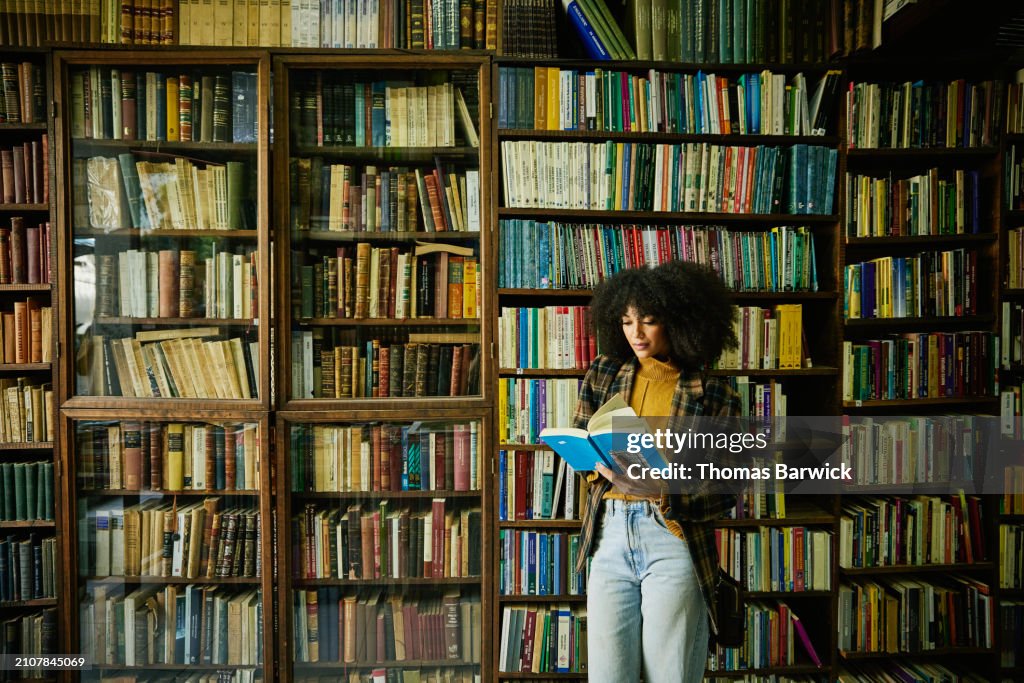 Wide shot woman reading book while browsing in antique bookstore