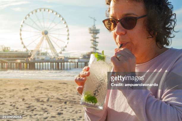 mature latin woman holding in her hands a fresh cocktail in a summer hot sunset time at scheveningen beach, the hague, the netherlands, europe. - scheveningen stock pictures, royalty-free photos & images