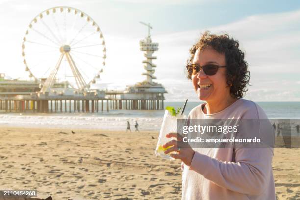 mature latin woman holding in her hands a fresh cocktail in a summer hot sunset time at scheveningen beach, the hague, the netherlands, europe. - scheveningen stock pictures, royalty-free photos & images