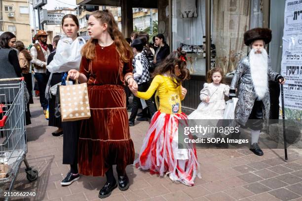 Children walk along the main street dressed in costumes; a boy, dressed as a Rabbi with a long white beard, holds the hand of a young girl dressed as...