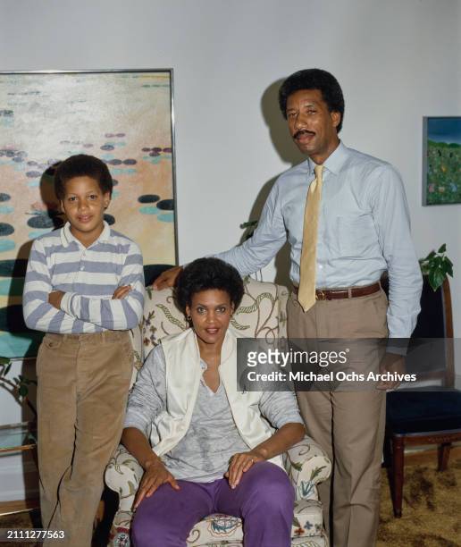 American journalist Charlayne Hunter-Gault, wearing a white waistcoat over a grey sweatshirt and purple sweatpants, sits in an armchair beside her...
