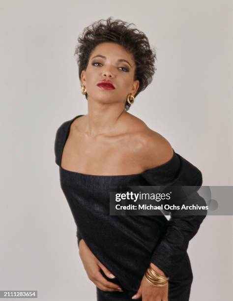 American actress Tisha Campbell, wearing a black minidress, gold hoop earrings and gold bangles, in a studio portrait session, against a white...