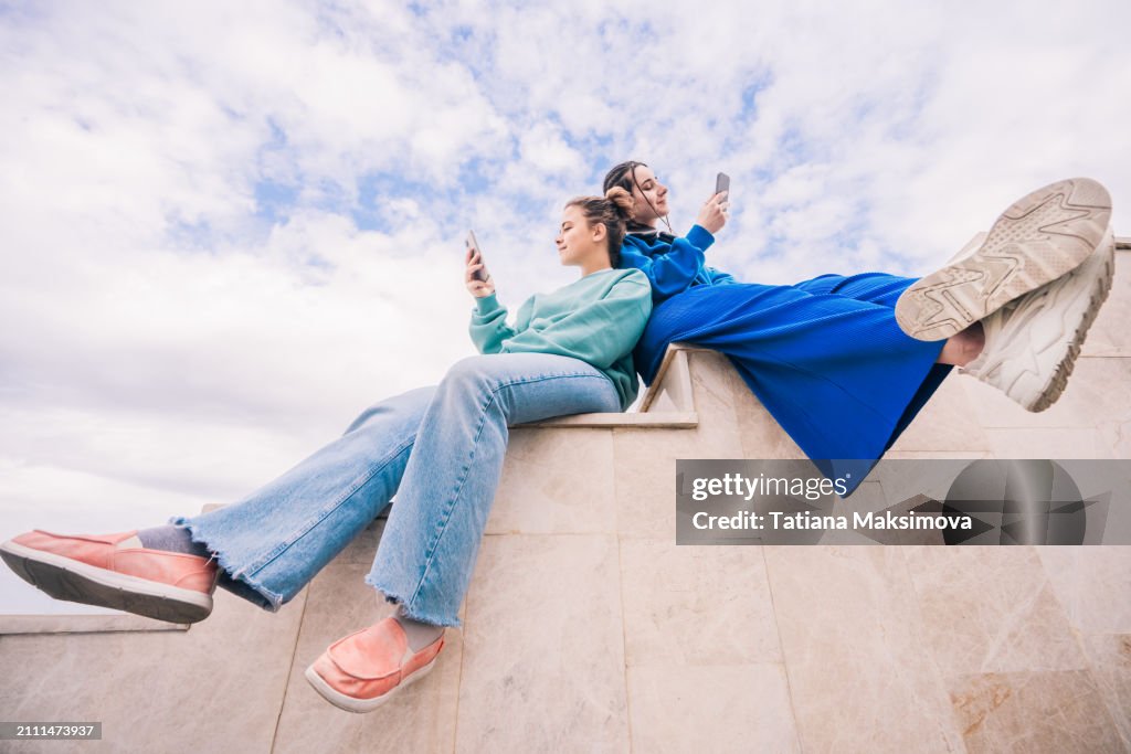 Two young female friends sitting with their phones on a staircase against the sky, view from below.