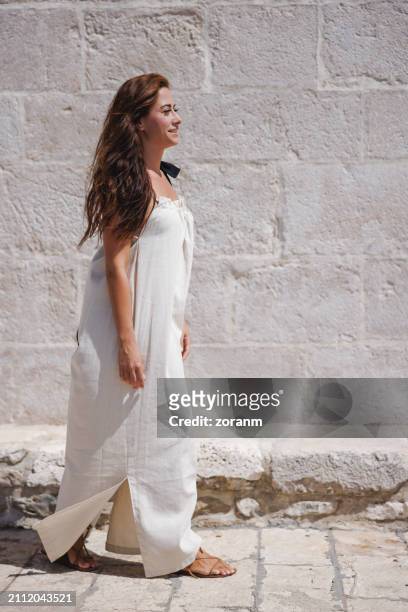 beautiful brunette walking on sunlight by stone wall, wearing long white sleeveless dress - sleeveless dress stock pictures, royalty-free photos & images
