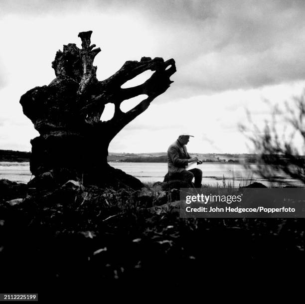 English artist Graham Sutherland sketches a rural scene as he sits on the exposed roots and trunk of a dead tree beside a river estuary near Milford...