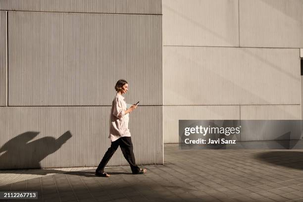femme d’affaires occupée et confiante marchant dans la rue de la ville tout en utilisant un téléphone portable dans la lumière du coucher du soleil - marches photos et images de collection