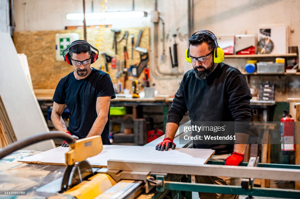 Carpenter cutting wood with electric saw near colleague at workshop