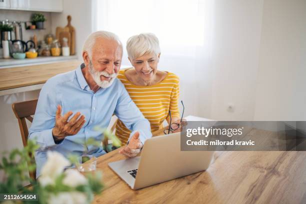 a senior couple is talking to family and friends on a video call via a laptop while sitting in the living room of their modern house - two people video conferencing stock pictures, royalty-free photos & images