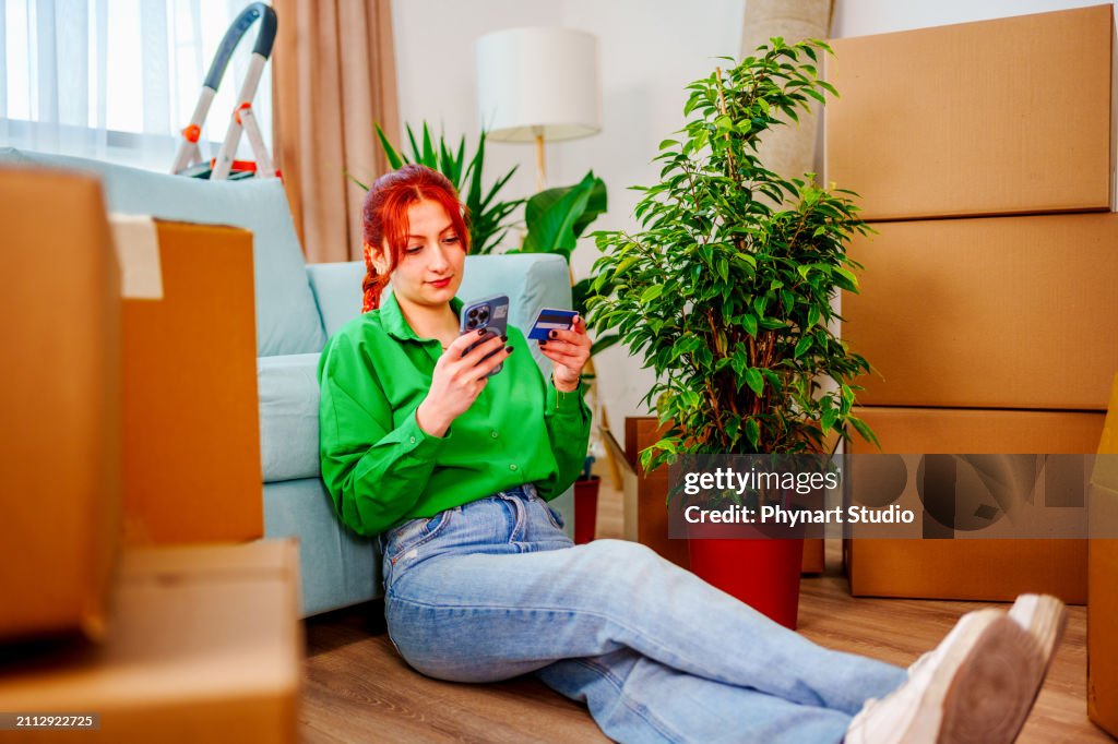Young woman moving into a new house is shopping and ordering with her mobile phone and credit card.