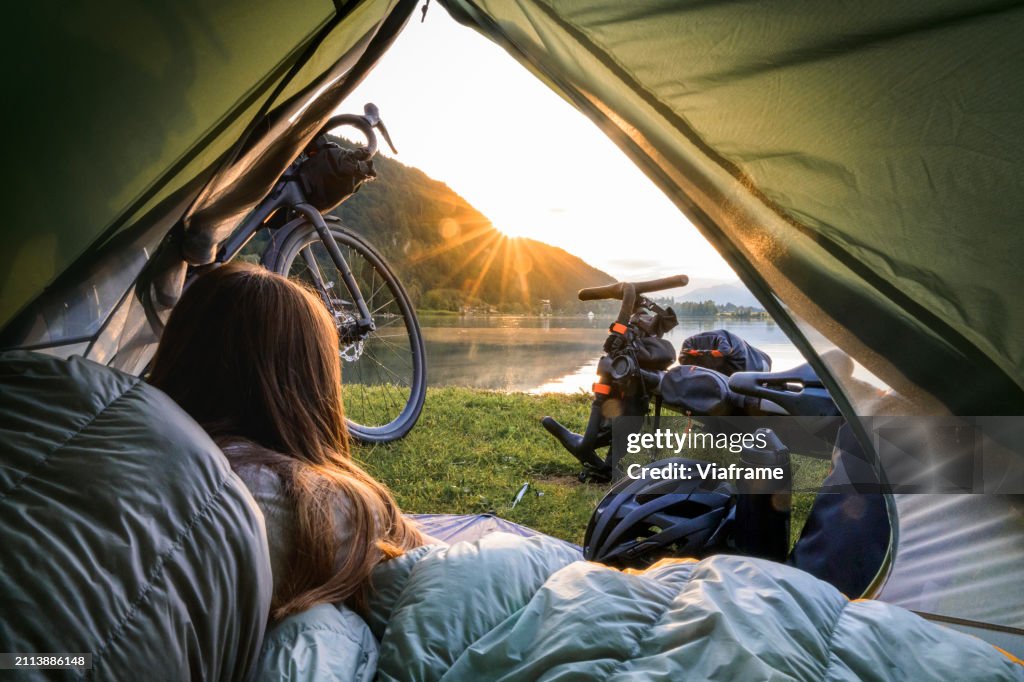 Young woman waking up in the early morning with a nice view out of her tent. She is traveling using a bike.