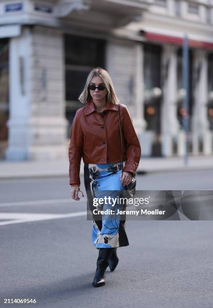 Karin Teigl seen wearing Miu Miu black oval sunglasses, Zara brown short leather jacket, Zara blue / white / black print pattern midi long skirt,...