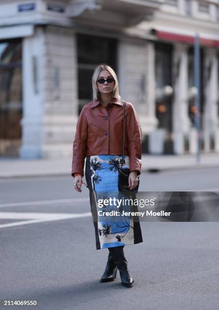 Karin Teigl seen wearing Miu Miu black oval sunglasses, Zara brown short leather jacket, Zara blue / white / black print pattern midi long skirt,...