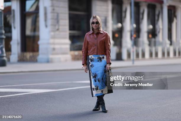 Karin Teigl seen wearing Miu Miu black oval sunglasses, Zara brown short leather jacket, Zara blue / white / black print pattern midi long skirt,...