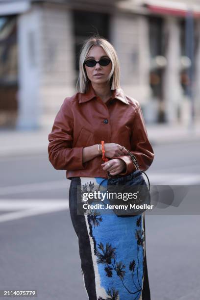 Karin Teigl seen wearing Miu Miu black oval sunglasses, Zara brown short leather jacket, Zara blue / white / black print pattern midi long skirt,...