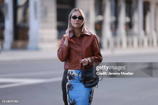 Karin Teigl seen wearing Miu Miu black oval sunglasses, Zara brown short leather jacket, Zara blue / white / black print pattern midi long skirt,...