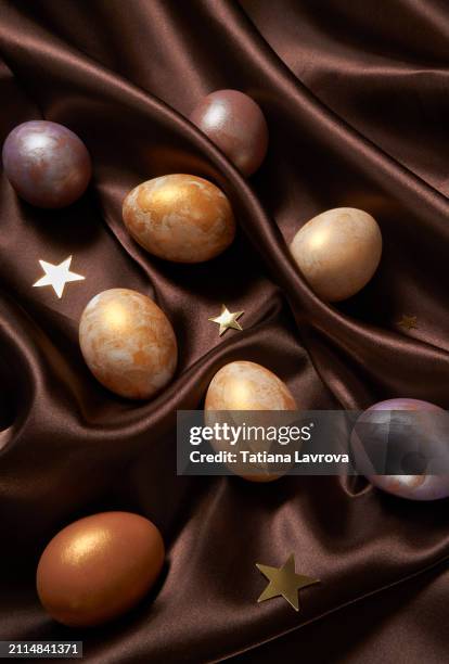 brown hued folded satin fabric background with elegant hand painted white, golden yellow  and violet easter eggs and star-shaped confetti. festive spring backdrop - star confetti white background stockfoto's en -beelden