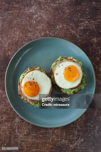 image of breakfast, blue-green plate of wholemeal, toasted bagel topped with mashed avocado, ham bacon rasher, sunny-side-up fried eggs, cracked peppercorn seasoning, wood grain background, elevated view, focus on foreground - fried egg stock pictures, royalty-free photos & images