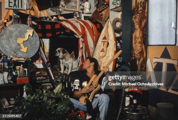 American comedian and actor Joe Piscopo, wearing a dark blue American football shirt with yellow and white flashes on the sleeves, sits holding an...