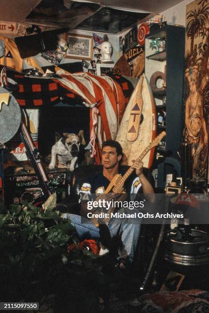 American comedian and actor Joe Piscopo, wearing a dark blue American football shirt with yellow and white flashes on the sleeves, sits holding an...