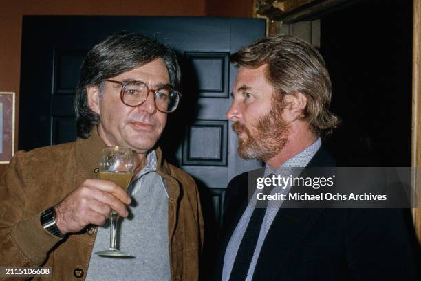 American film director Richard Donner, wearing a brown suede jacket over a grey sweater and a striped shirt, holding a drinking glass as he stands...