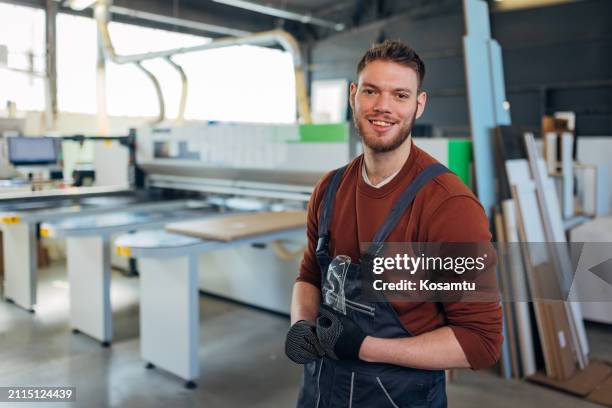 a smiling worker in coveralls and protective gloves looks directly at the camera while standing in the workshop - overall stockfoto's en -beelden