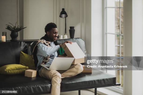 joyful black man unpacking shopping box with new clothes sitting on couch at home. - commercial event stock pictures, royalty-free photos & images