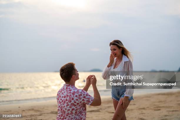 romantic beach wedding proposal: caucasian man surprises girlfriend with a sunset engagement by the ocean - fiançailles photos et images de collection