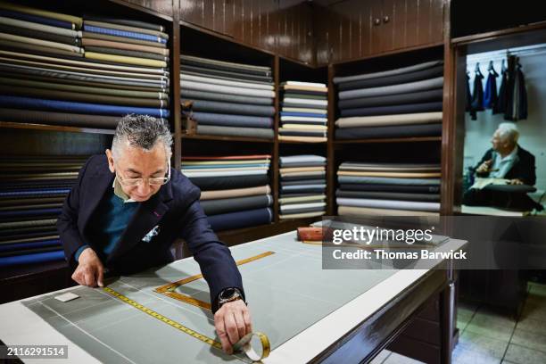 medium wide shot tailor measuring fabric for suit at table in workshop - tailor stock pictures, royalty-free photos & images