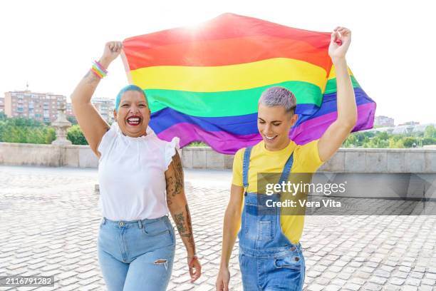 front portrait of homosexual couple holding a rainbow flag outdoors with happy expression - lgbtq  female fotografías e imágenes de stock