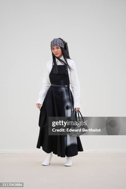 Yoyo Cao wears a Dior monogram printed scarf / bandanna over the head, circular earrings, a white shirt, a thin belt, black overalls / dungarees worn...