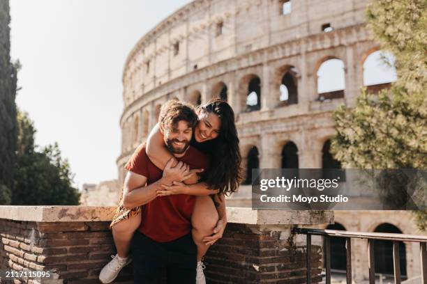 happy bearded man and girlfriend playing piggyback whilst on vacation, colosseum in background - rome italy stock pictures, royalty-free photos & images