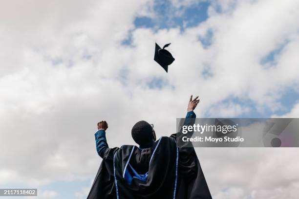 low angle view of man in graduating gown throwing mortarboard into air on graduation day - graduation stock pictures, royalty-free photos & images