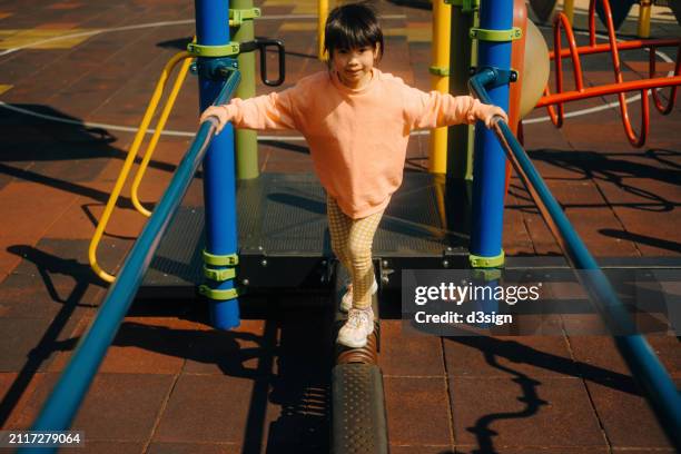 portrait of a lovely little asian girl smiling at the camera while playing joyfully at the balancing equipments in an outdoor playground. enjoying outdoor fun time at the park. carefree and freedom concept - playground balance beam stock pictures, royalty-free photos & images