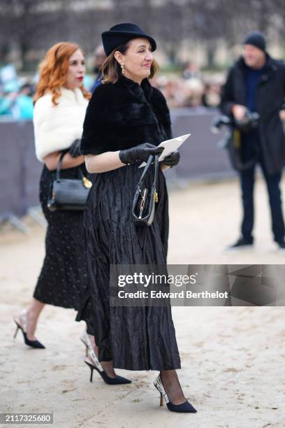 Guest wears a hat, earrings, a black fluffy fur coat, black leather gloves, a black gathered skirt , high heels pointed shoes, outside Dior, during...