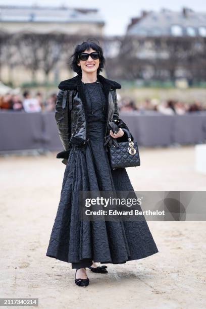 Guest wears sunglasses, a black shiny leather aviator jacket with fluffy inner lining , a black texture gathered / pleated long dress , a Lady Dior...