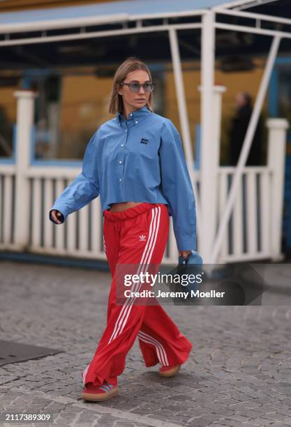 Karin Teigl seen wearing Rhude grey sunglasses, Miu Miu blue cotton cropped buttoned shirt, Adidas red long wide leg track pants, Prada Re-edition...