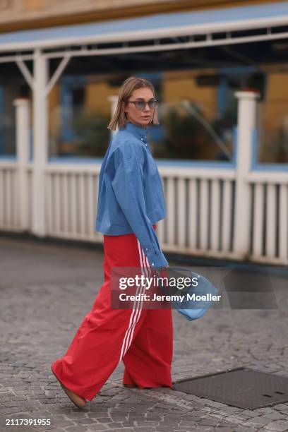 Karin Teigl seen wearing Rhude grey sunglasses, Miu Miu blue cotton cropped buttoned shirt, Adidas red long wide leg track pants, Prada Re-edition...