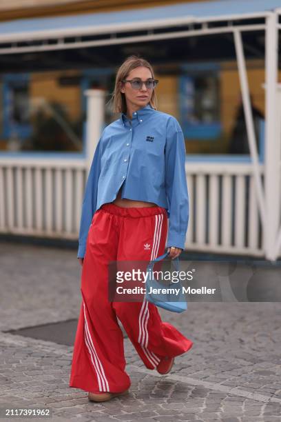Karin Teigl seen wearing Rhude grey sunglasses, Miu Miu blue cotton cropped buttoned shirt, Adidas red long wide leg track pants, Prada Re-edition...
