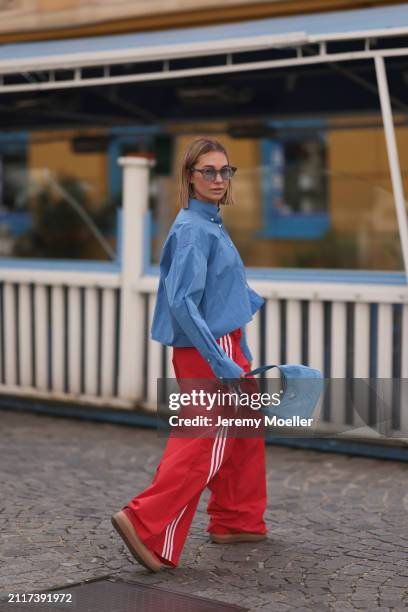 Karin Teigl seen wearing Rhude grey sunglasses, Miu Miu blue cotton cropped buttoned shirt, Adidas red long wide leg track pants, Prada Re-edition...