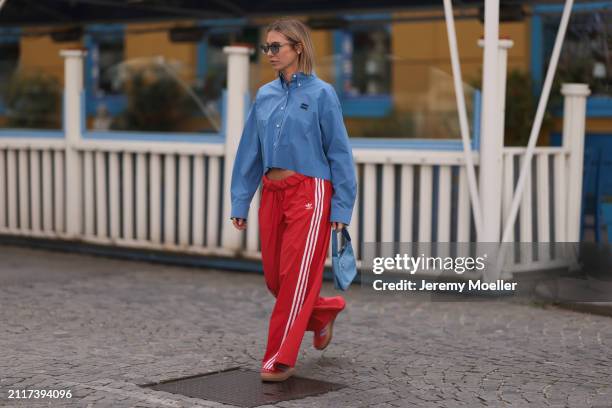 Karin Teigl seen wearing Rhude grey sunglasses, Miu Miu blue cotton cropped buttoned shirt, Adidas red long wide leg track pants, Prada Re-edition...