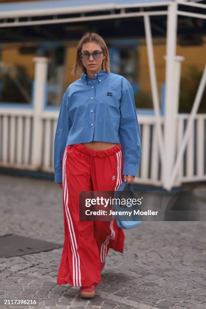 Karin Teigl seen wearing Rhude grey sunglasses, Miu Miu blue cotton cropped buttoned shirt, Adidas red long wide leg track pants, Prada Re-edition...