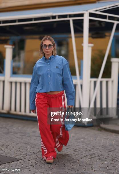 Karin Teigl seen wearing Rhude grey sunglasses, Miu Miu blue cotton cropped buttoned shirt, Adidas red long wide leg track pants, Prada Re-edition...