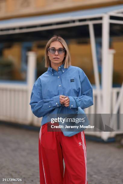 Karin Teigl seen wearing Rhude grey sunglasses, Miu Miu blue cotton cropped buttoned shirt, Adidas red long wide leg track pants, Prada Re-edition...