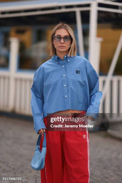 Karin Teigl seen wearing Rhude grey sunglasses, Miu Miu blue cotton cropped buttoned shirt, Adidas red long wide leg track pants, Prada Re-edition...