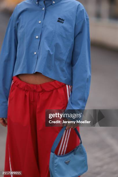 Karin Teigl seen wearing Miu Miu blue cotton cropped buttoned shirt, Adidas red long wide leg track pants, Prada Re-edition blue nylon bag and...