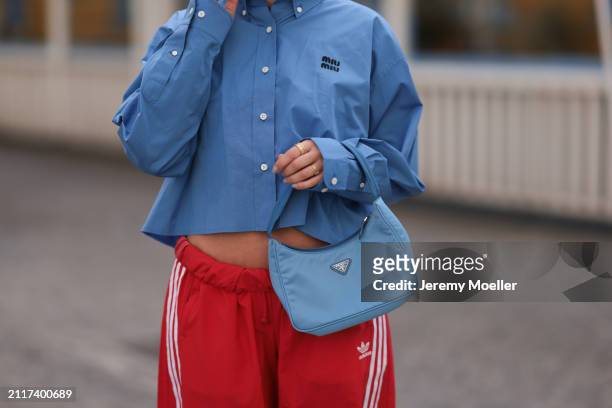 Karin Teigl seen wearing Miu Miu blue cotton cropped buttoned shirt, Adidas red long wide leg track pants, Prada Re-edition blue nylon bag and...